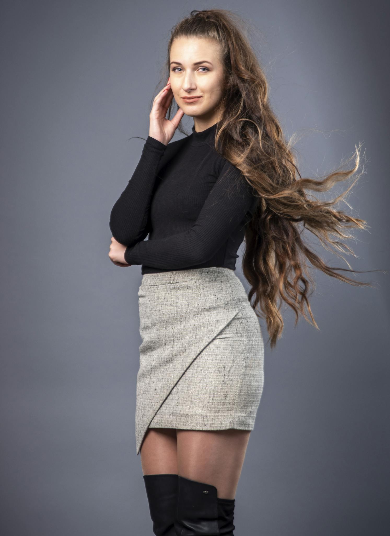 Elegant woman posing in a studio with long hair and fashionable attire. Black sweater with gray skirt.